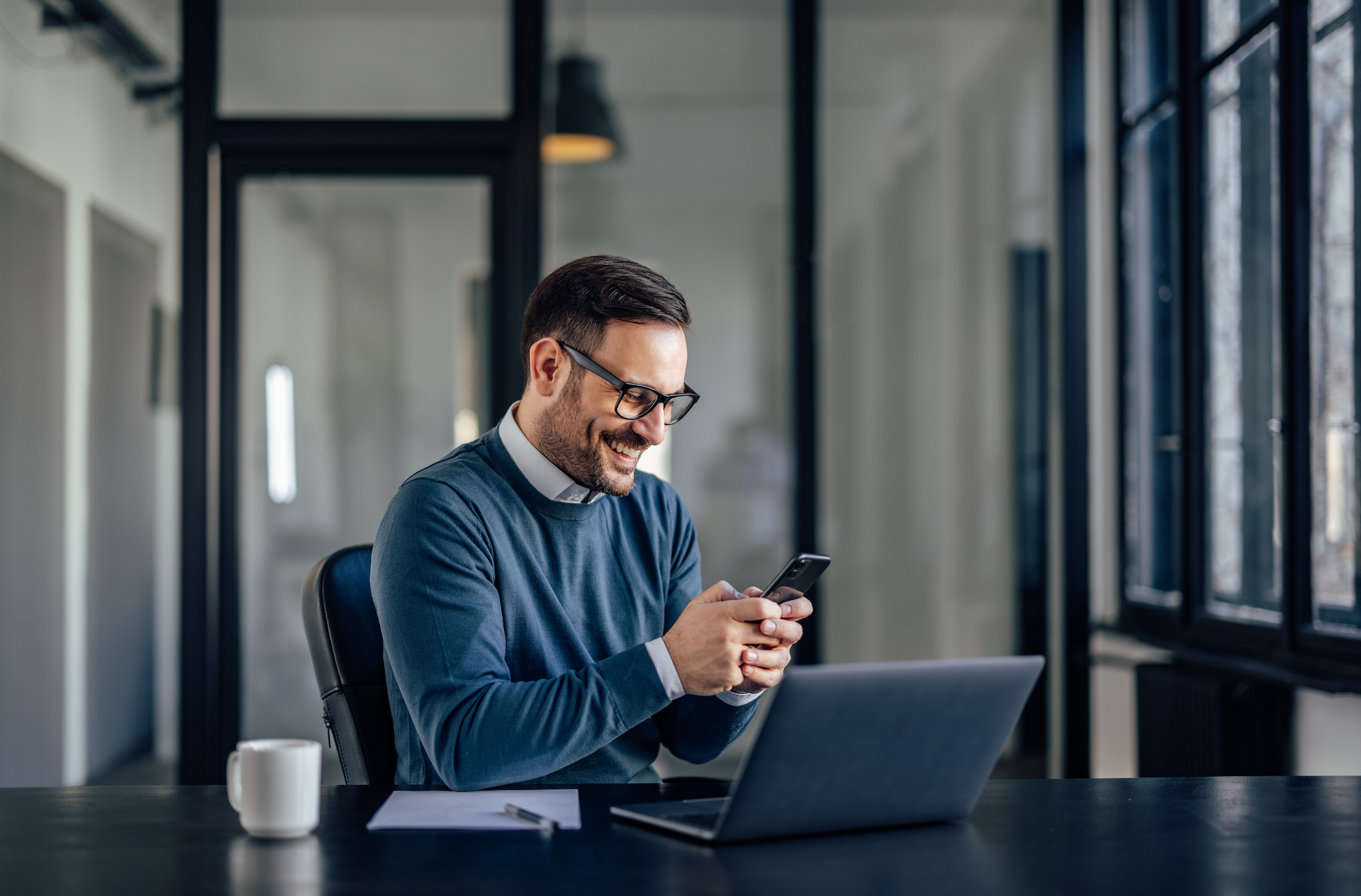 A man is sitting at a desk using a laptop and a cell phone.