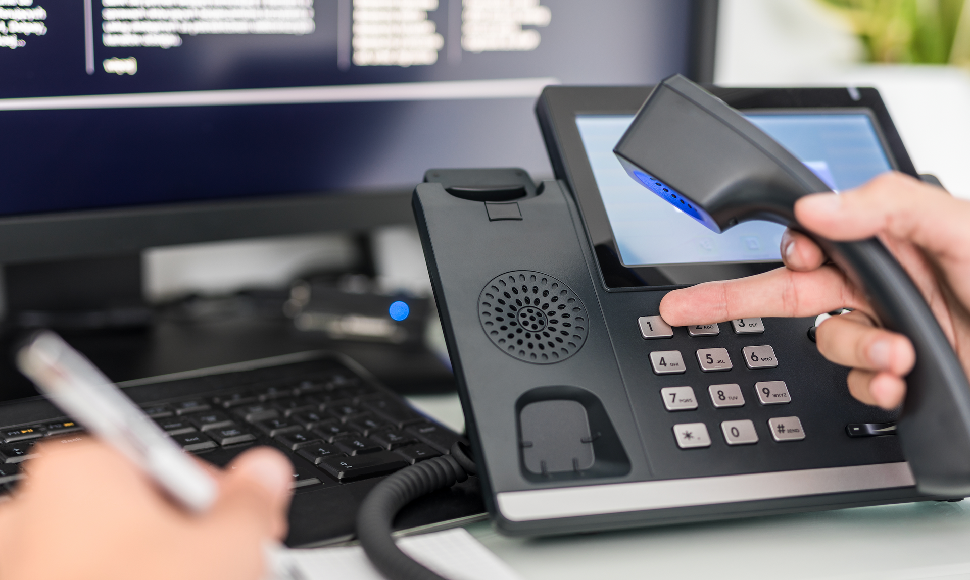 A person is holding a telephone in front of a computer.
