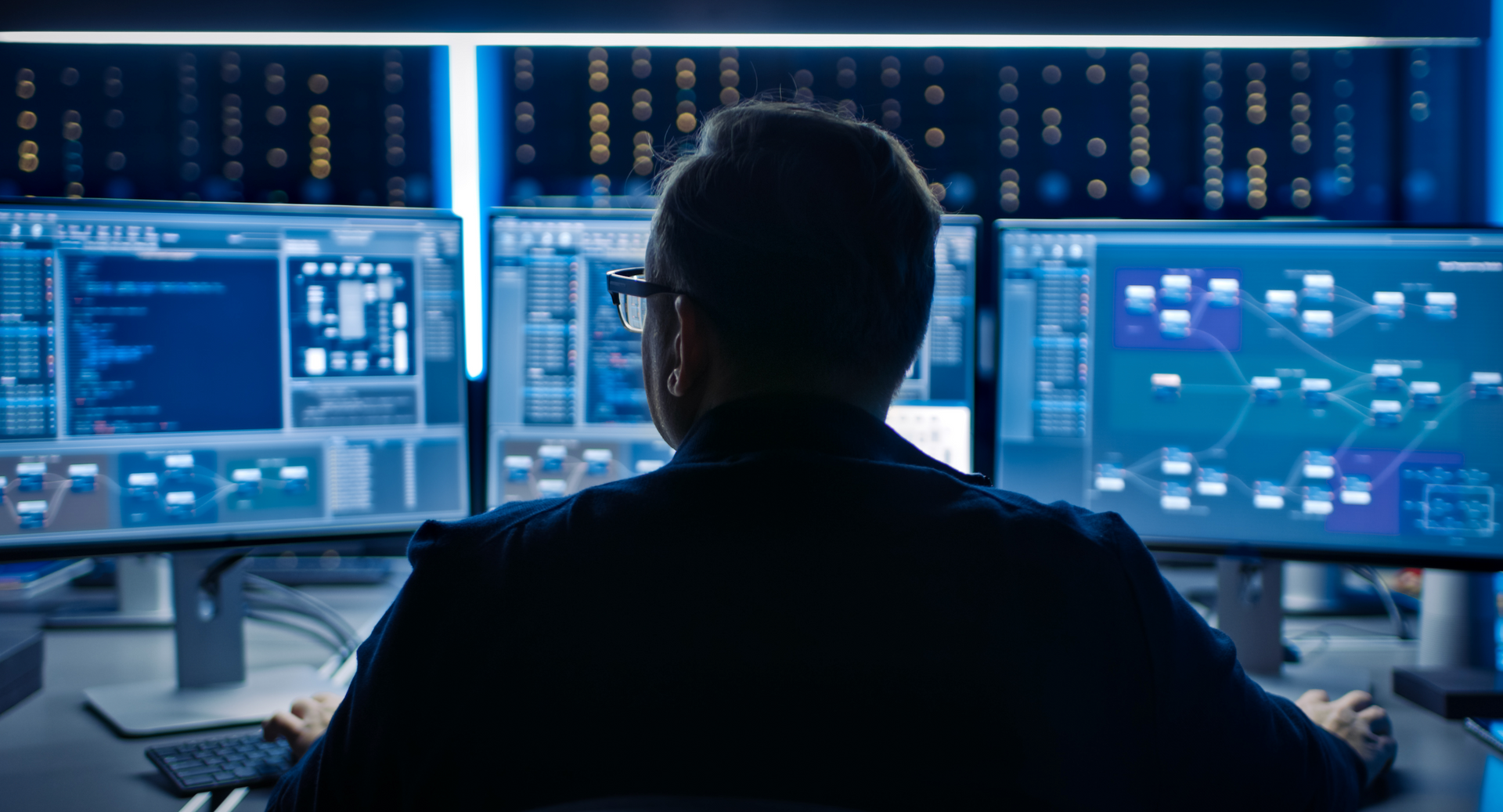 A man is sitting in front of three computer monitors in a dark room.