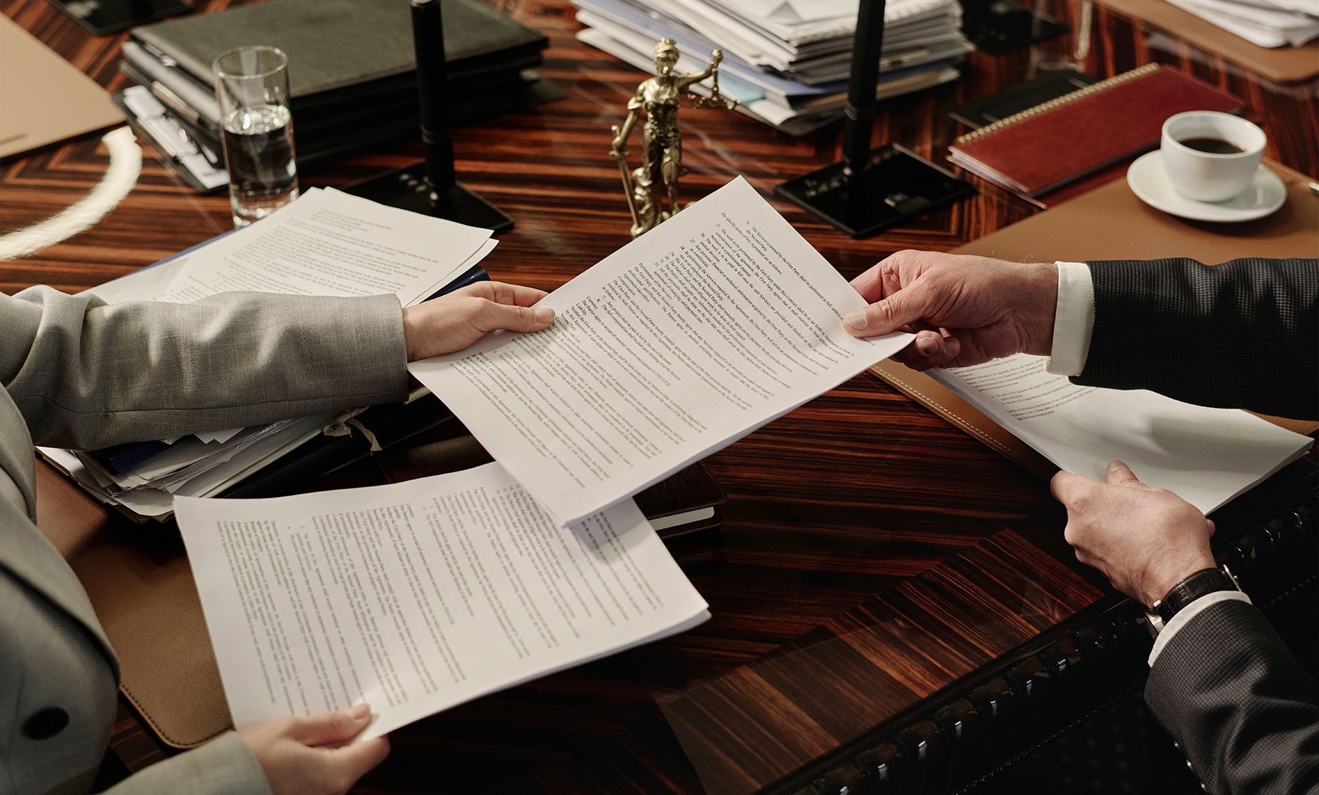 Hands exchanging documents at a wooden table, papers and coffee visible.
