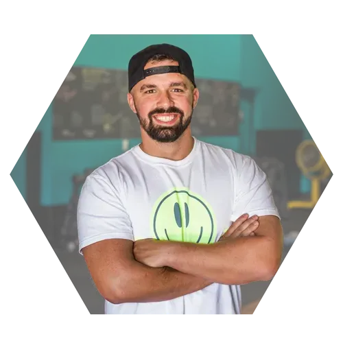 Man with crossed arms, smiling, wearing a baseball cap and a white tee with a green smiley face in a gym.