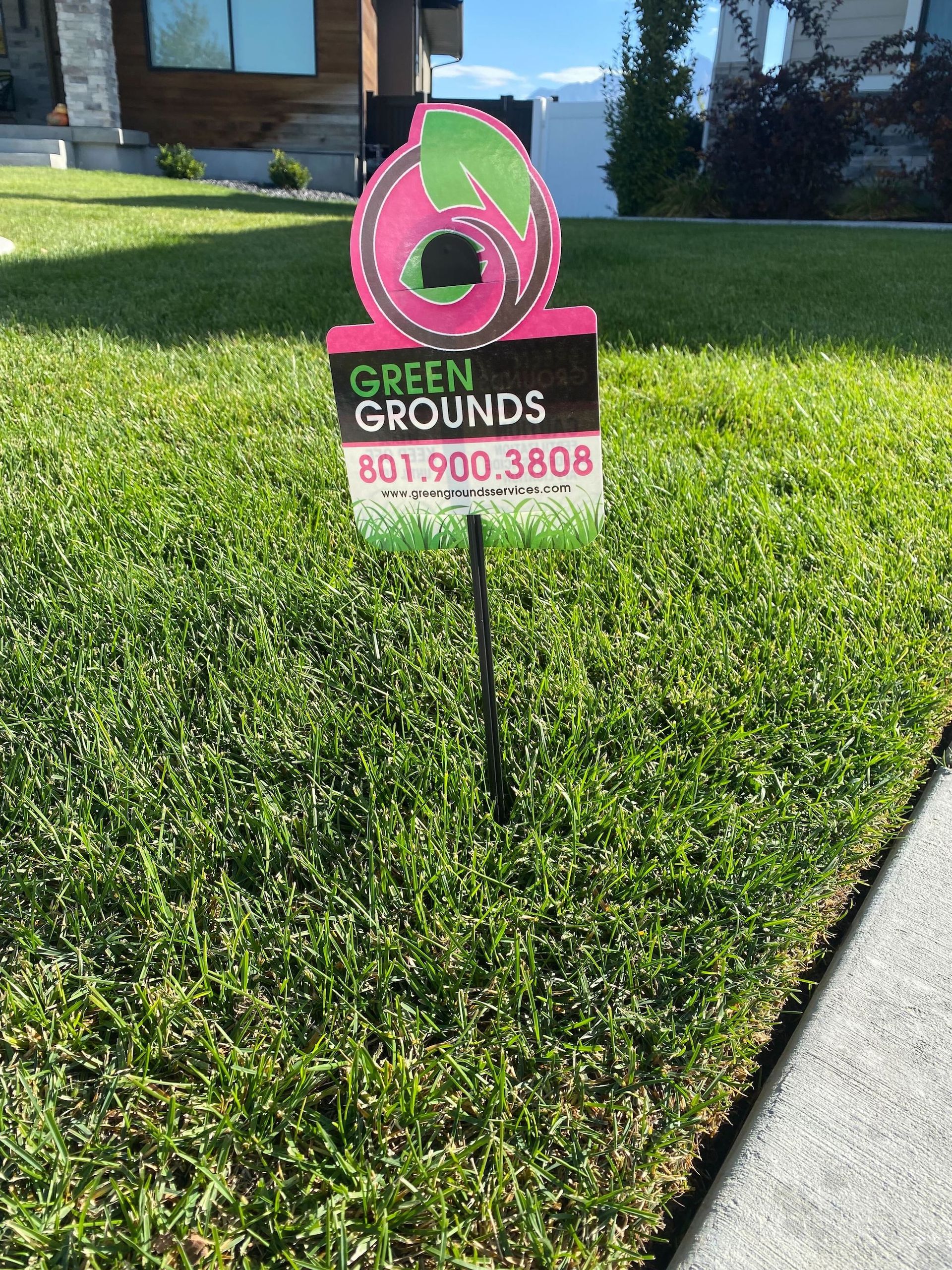 A sign for green grounds is sitting in the grass in front of a house.