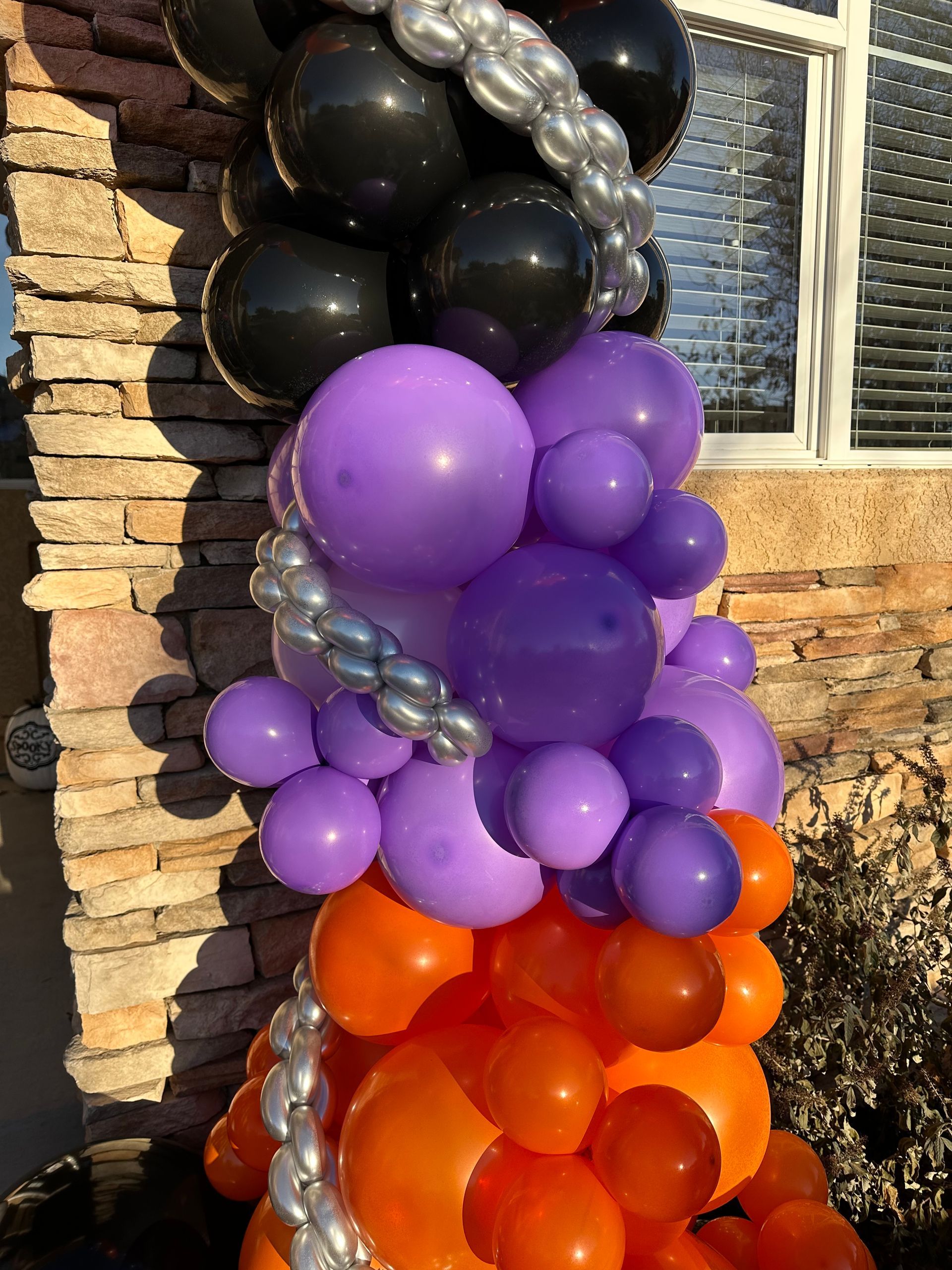 Balloon column in black, silver, purple, and orange against a brick wall and window.