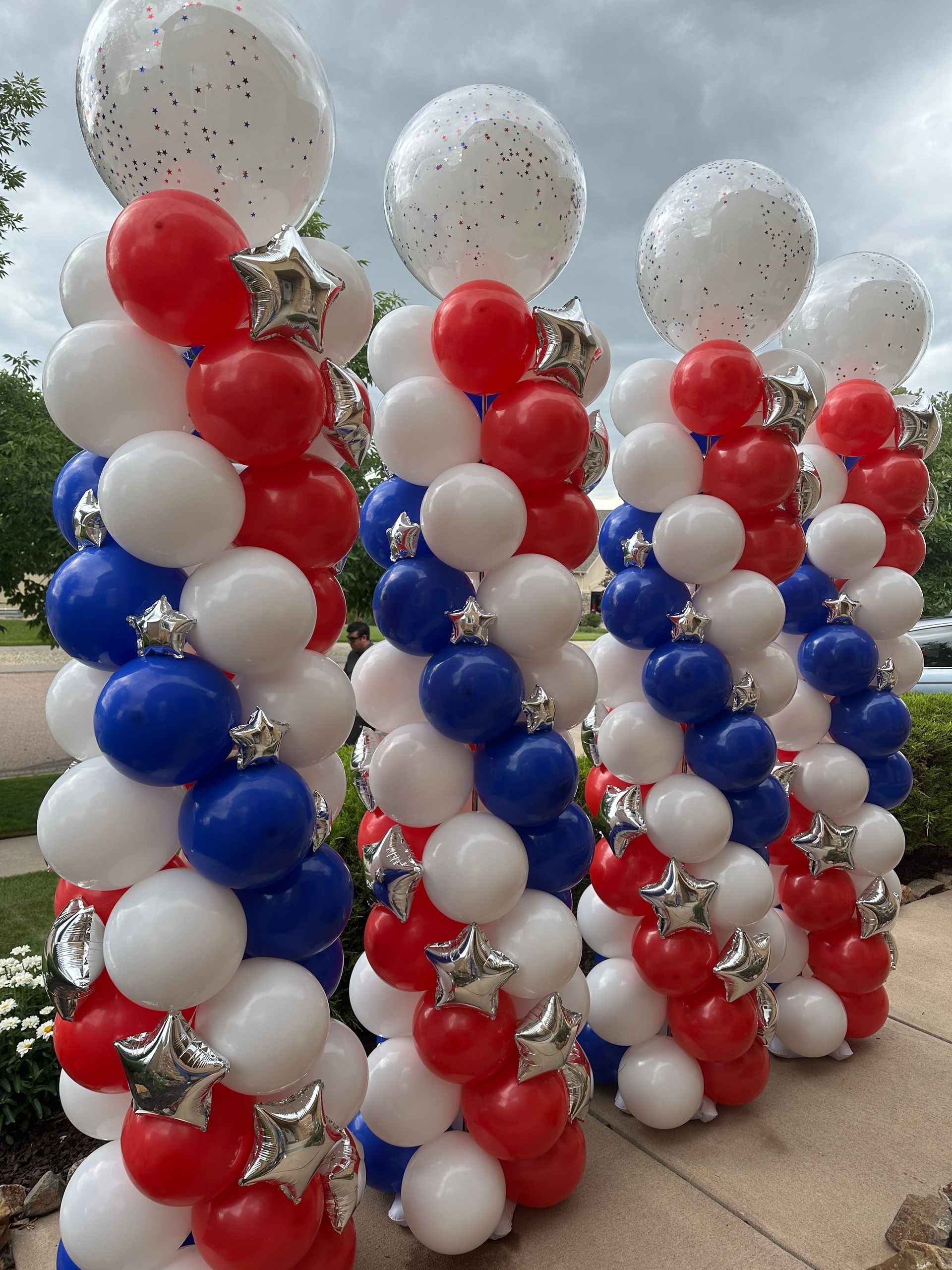 Red, white, and blue balloon columns topped with white confetti balloons, silver stars. Outdoors, likely a celebration.