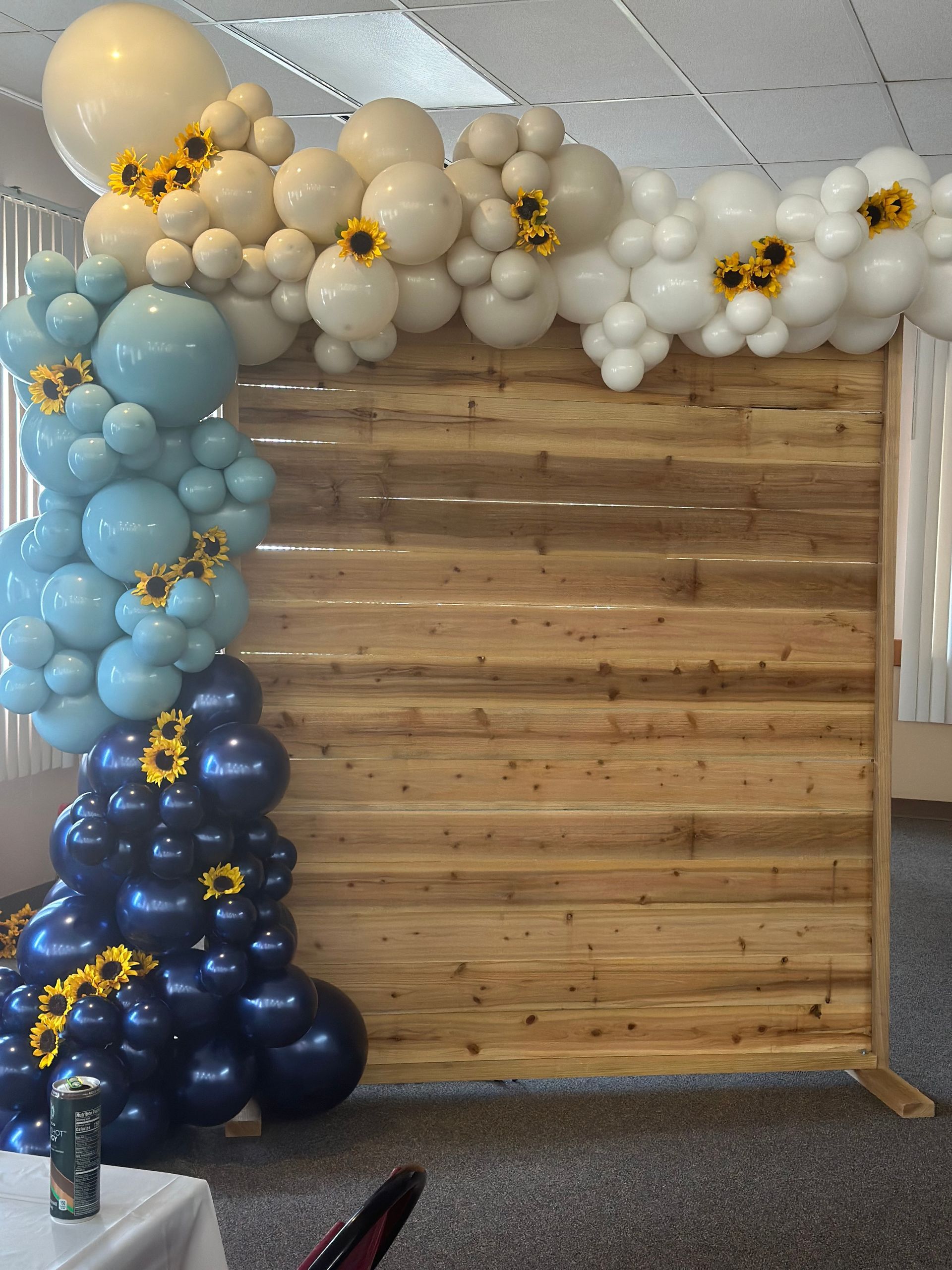 Balloon arch in blues, whites, and yellows with sunflowers, set against a wooden backdrop.