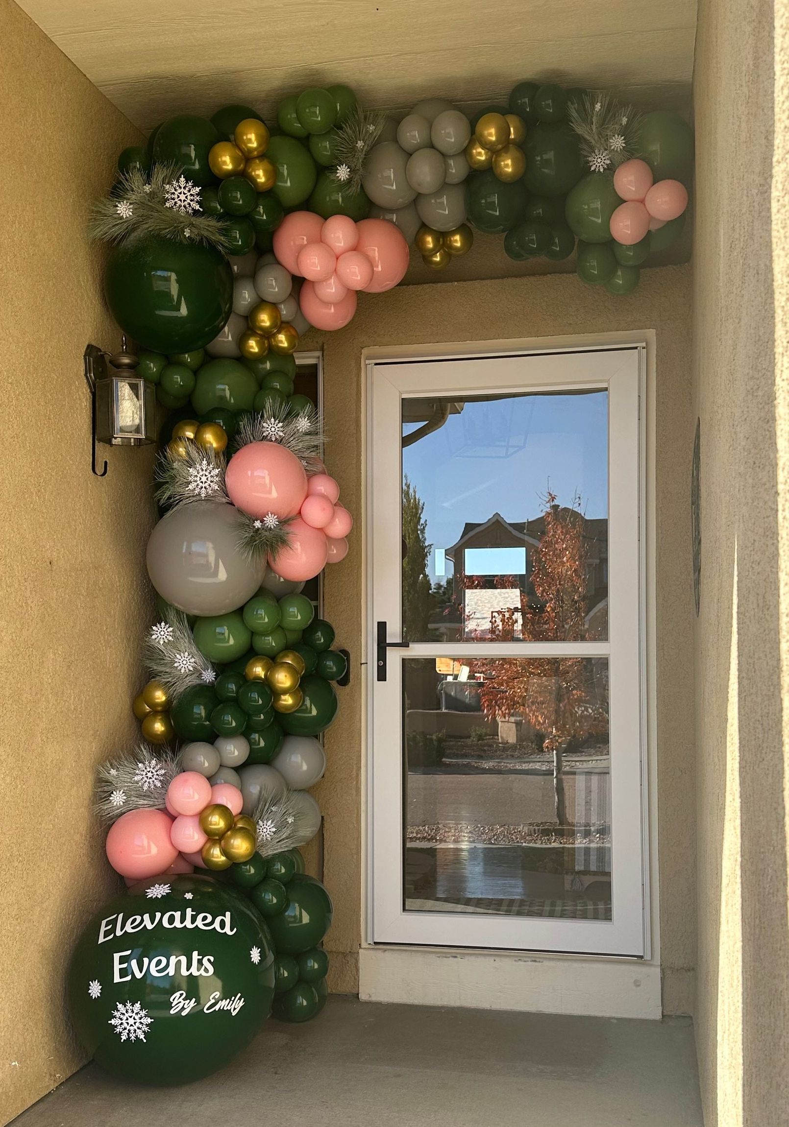 Arch of green, gold, pink, and grey balloons frames a white door with a reflective glass panel.
