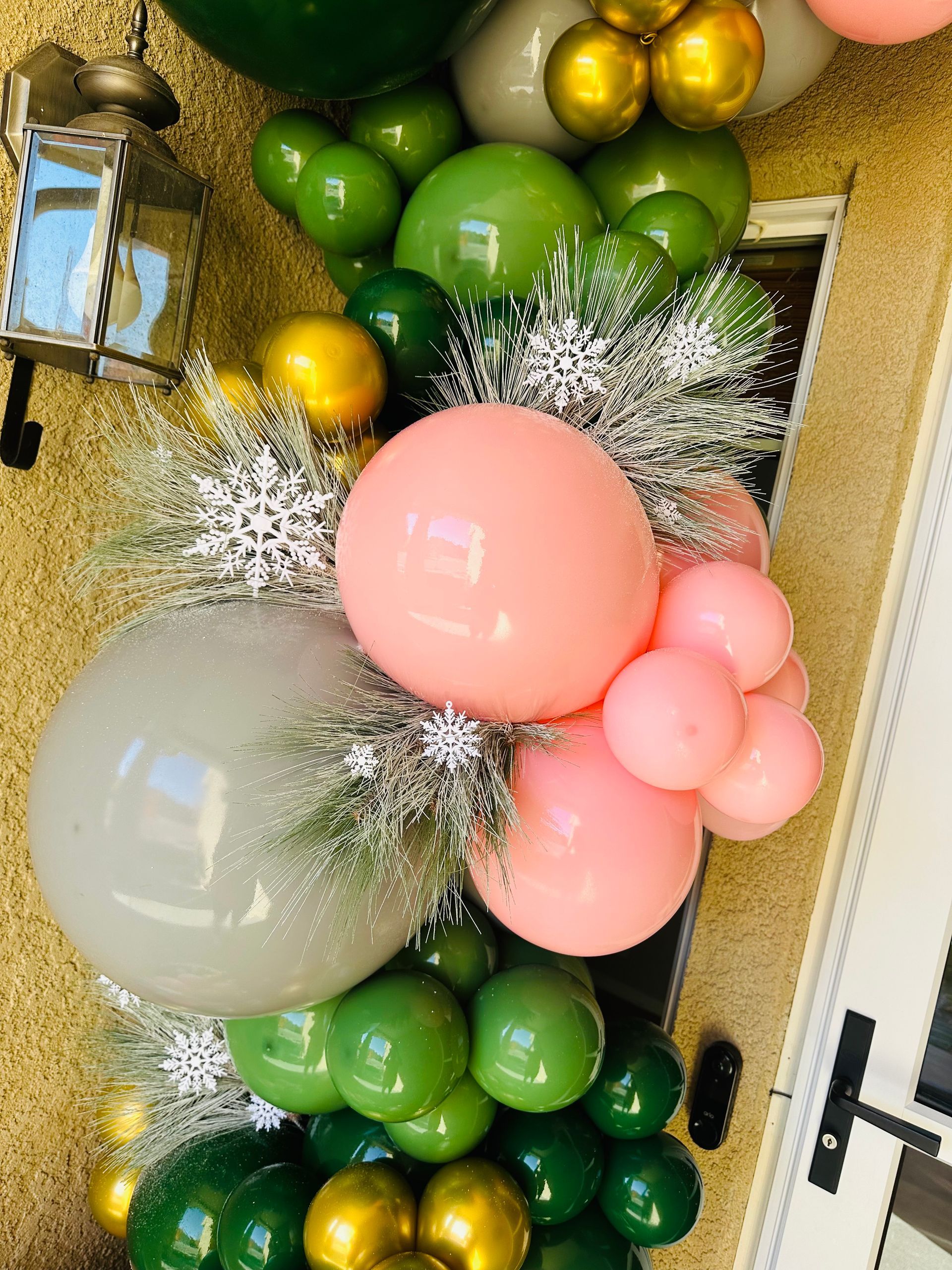 Balloon garland in shades of green, gold, pink, and gray with silver accents, against a stucco wall.