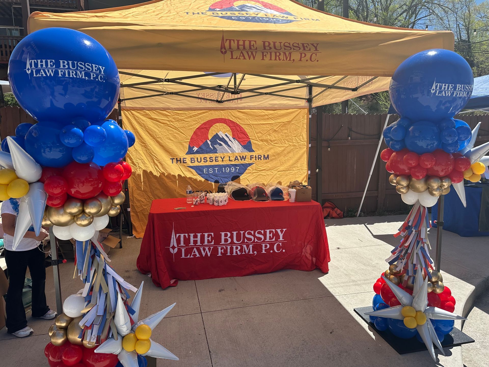 Law firm booth with red, white, and blue balloon decorations, including a tent and table with logos.