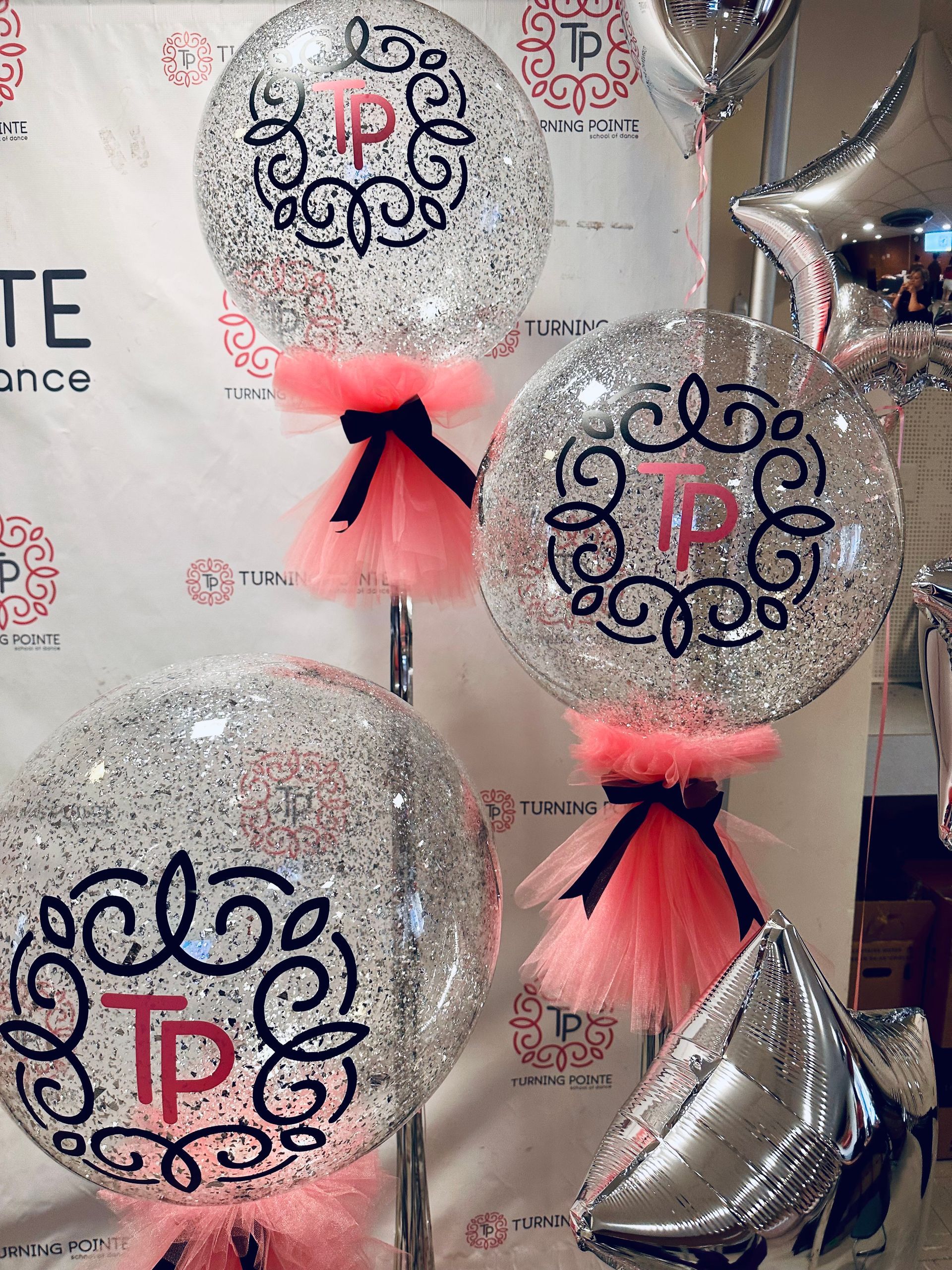 Clear balloons with silver glitter and pink tulle, featuring a logo, at an event.