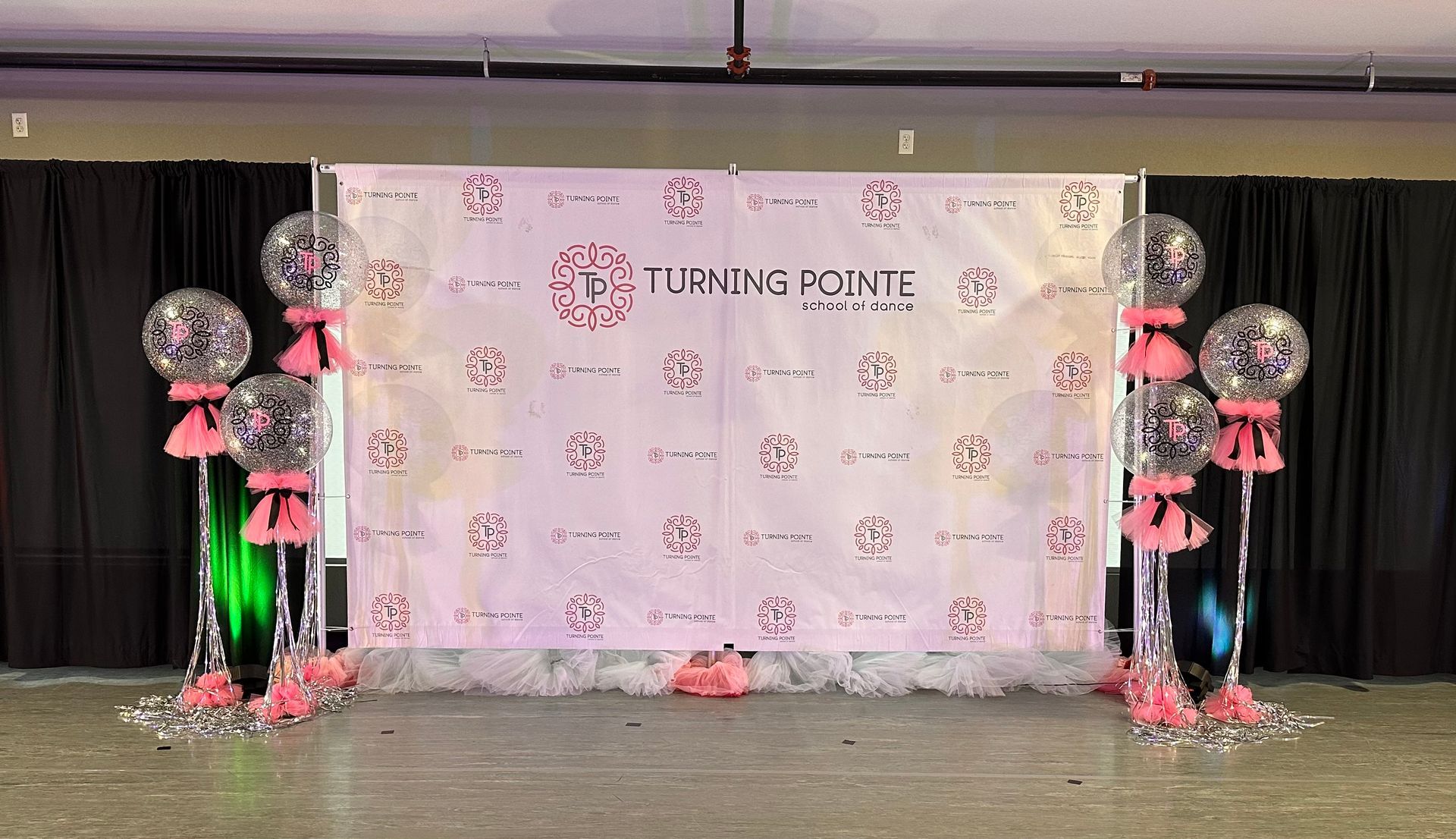 Event backdrop with pink logo and balloons; black curtains in a venue.