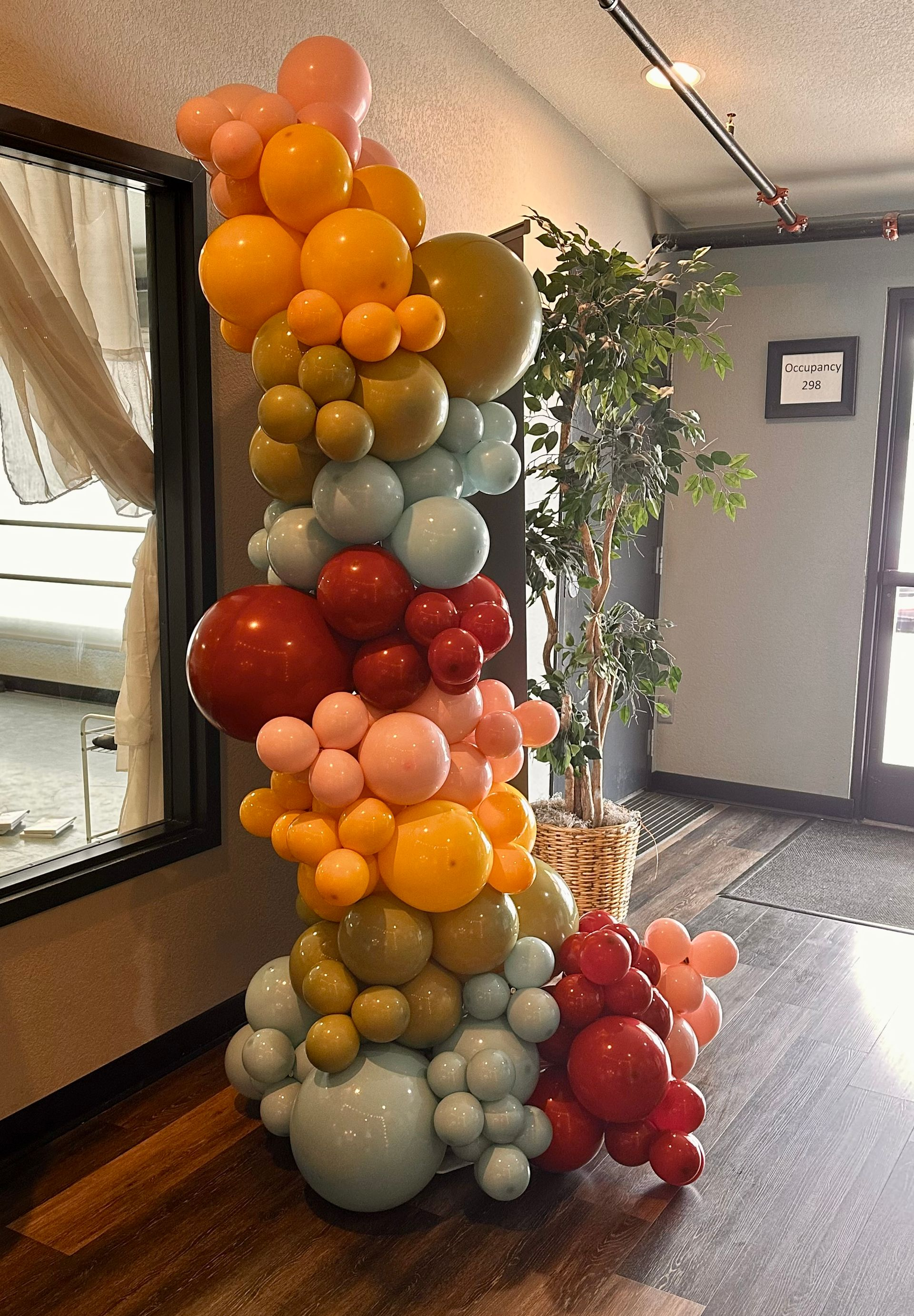 Colorful balloon column with various sizes and hues against a backdrop of a mirror, a plant, and a door.