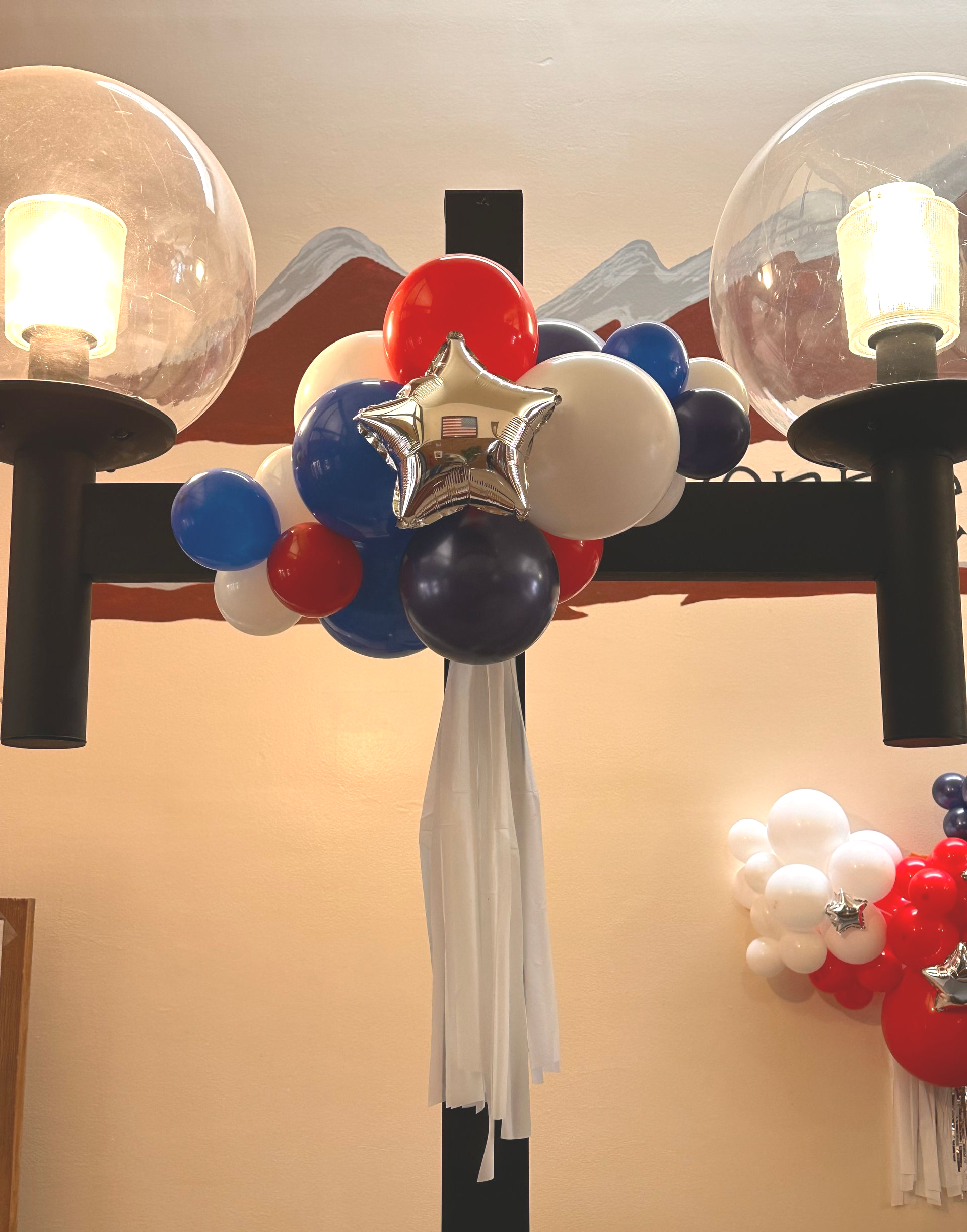 Balloons in red, white, and blue, with a star-shaped balloon, attached to a black structure under glass light fixtures.