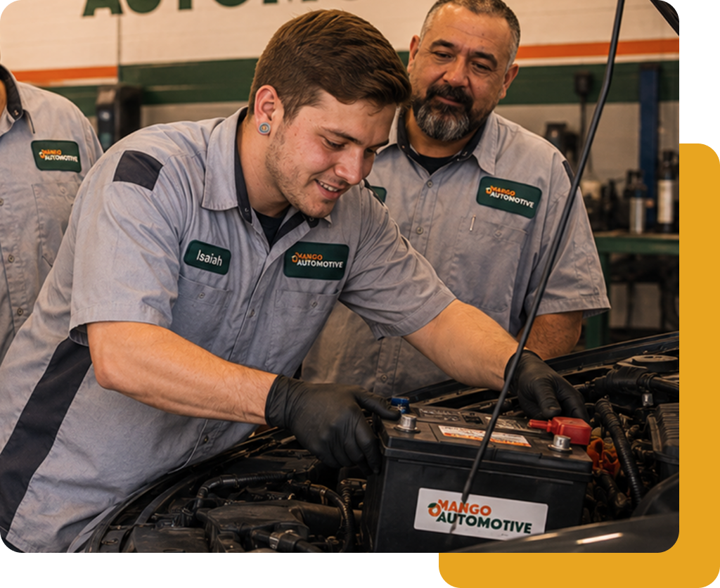 Two mechanics in gray uniforms work on a car engine in a garage, one smiling.