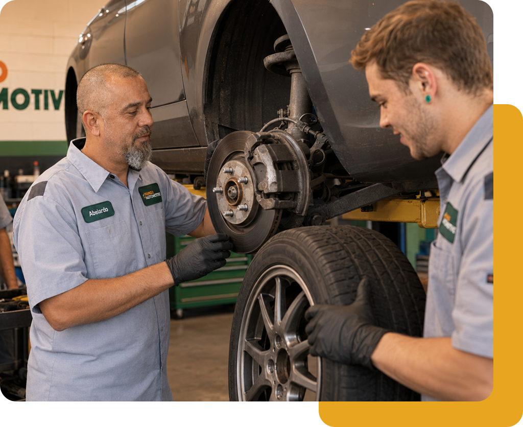 Two mechanics in gray uniforms inspect a car wheel and brake assembly in a garage.