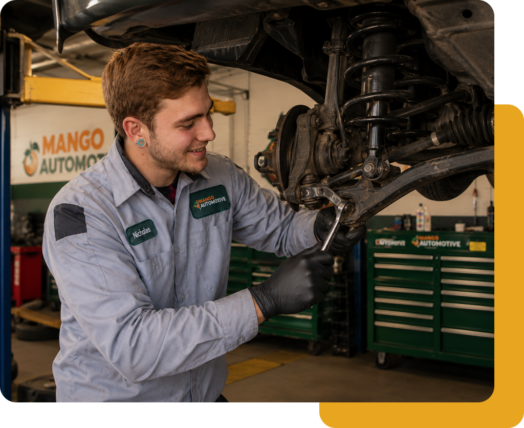 Mechanic in gray uniform working under a vehicle in an auto repair shop with green tool cabinets.