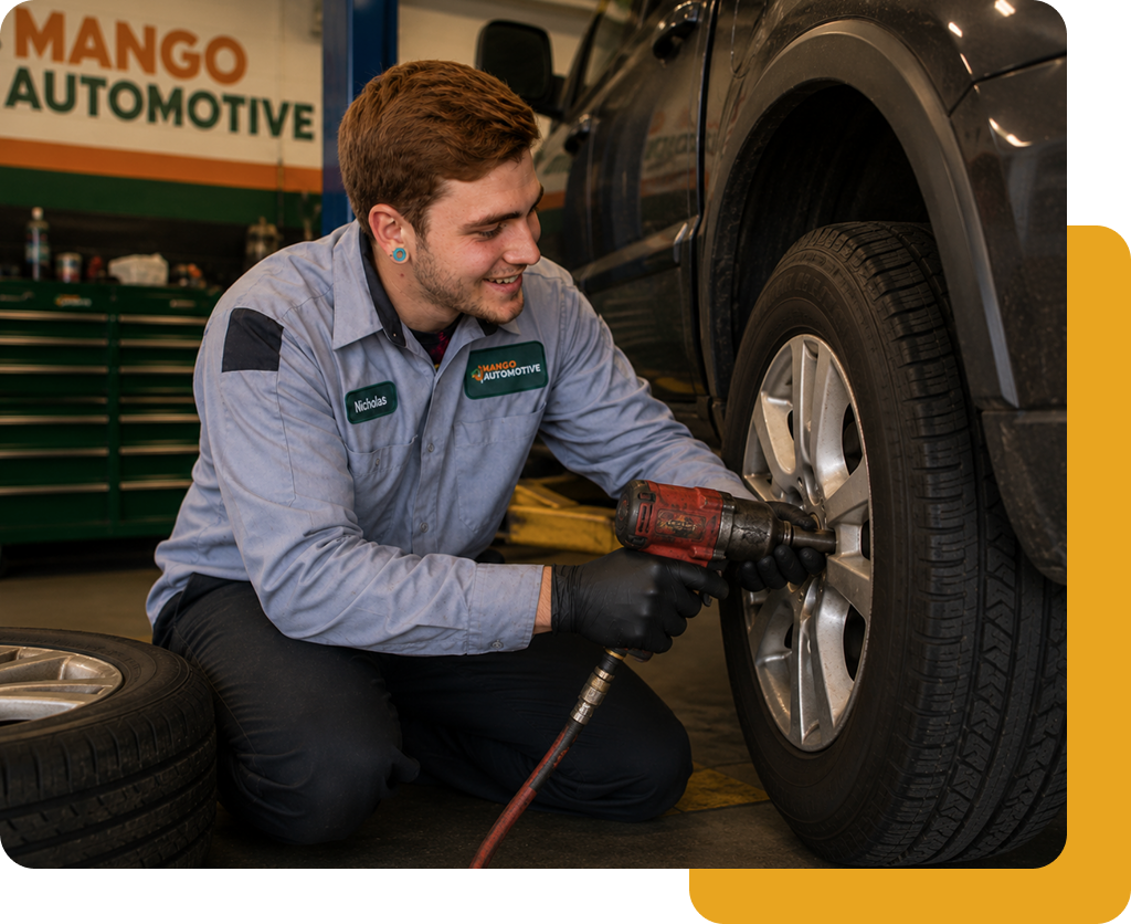 Mechanic tightening a car wheel with a power tool in an auto shop, next to a stack of tires.