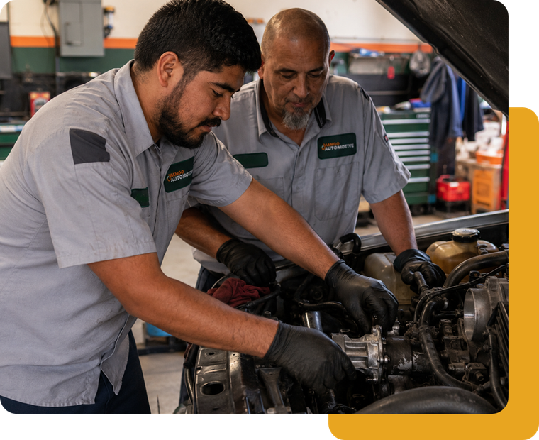 Two mechanics in gray uniforms inspect a car engine in a workshop with the hood open.