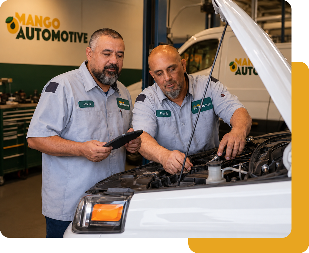 Two mechanics inspecting a white car engine in an auto shop, with a Mango Automotive sign in back.
