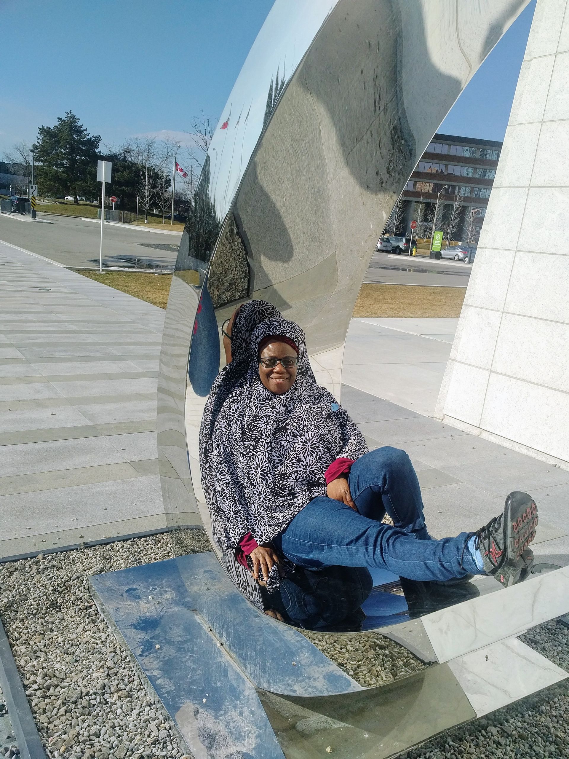 Person seated against a modern sculpture, wearing a patterned coat, jeans, and glasses, outdoors.