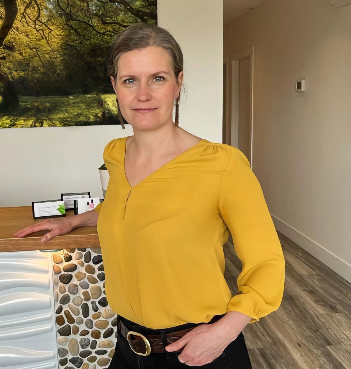 A woman with graying hair wearing a yellow blouse stands at a reception desk with a stone front and wooden countertop.