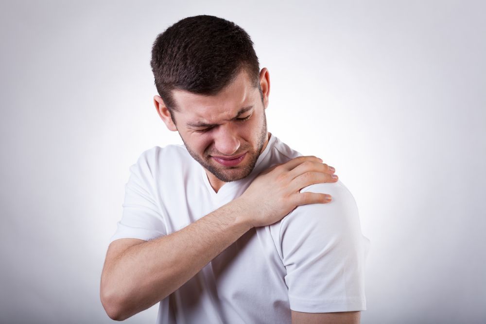 Man in a white t-shirt clutching his shoulder, indicating pain, with a pained expression.