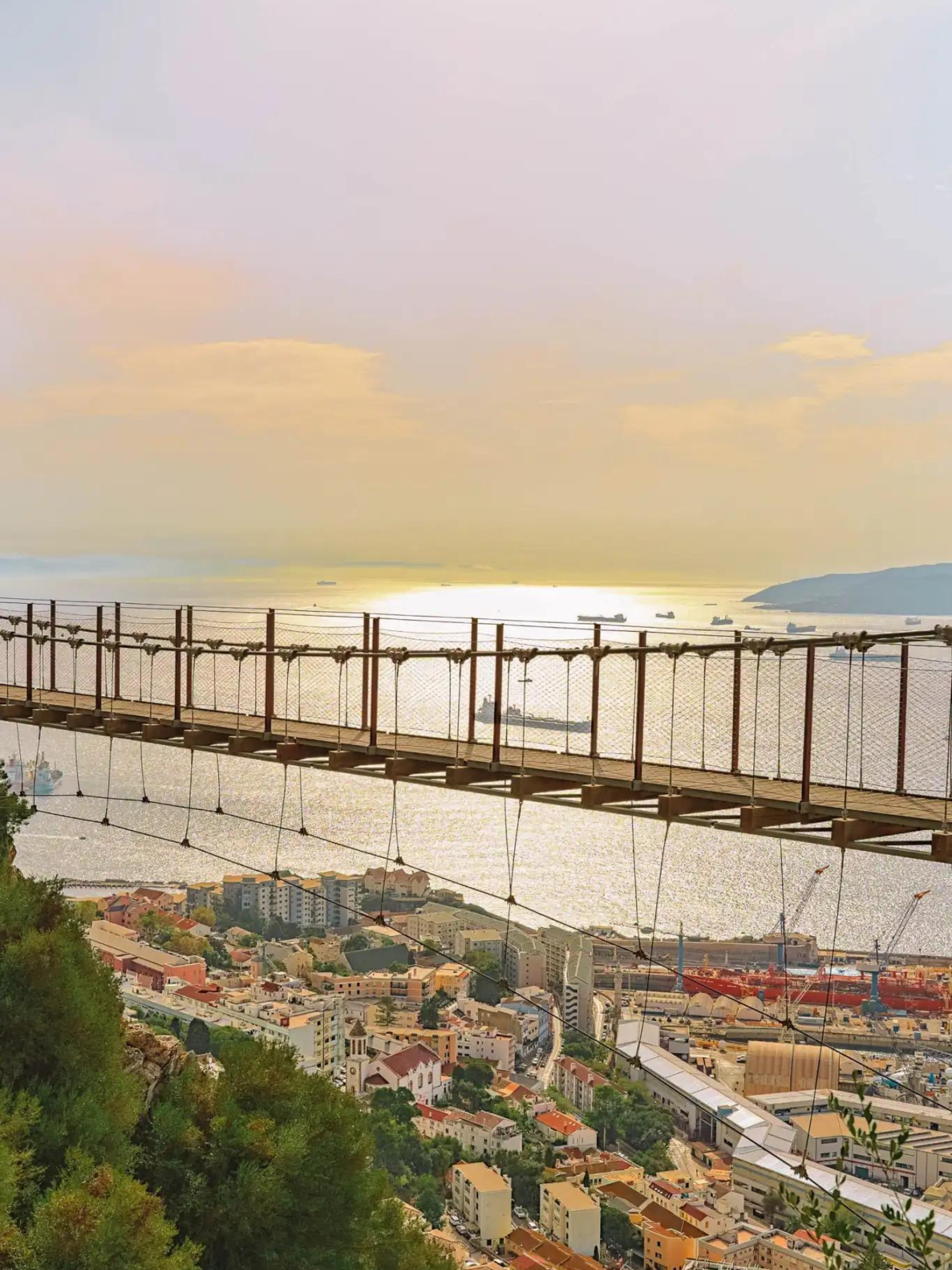 Suspension bridge over a coastal city at sunset, with water, hills, and a warm pastel sky