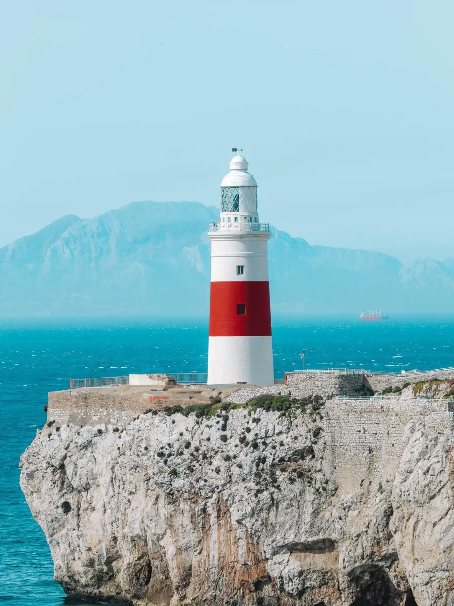 Red-and-white lighthouse on a rocky cliff above blue sea with mountains in the background