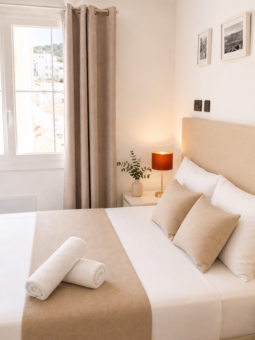 Bright bedroom with white bed, colorful pillows, nightstand, lamp, mirror, and window with curtains