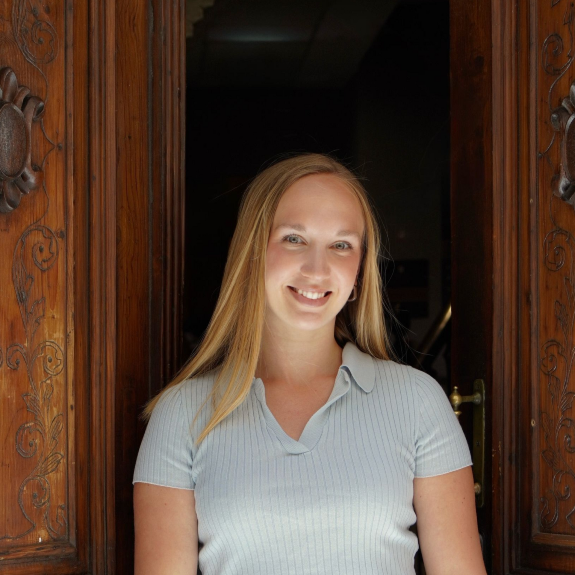 Smiling woman in a light blue shirt and brown skirt standing in a wooden doorway