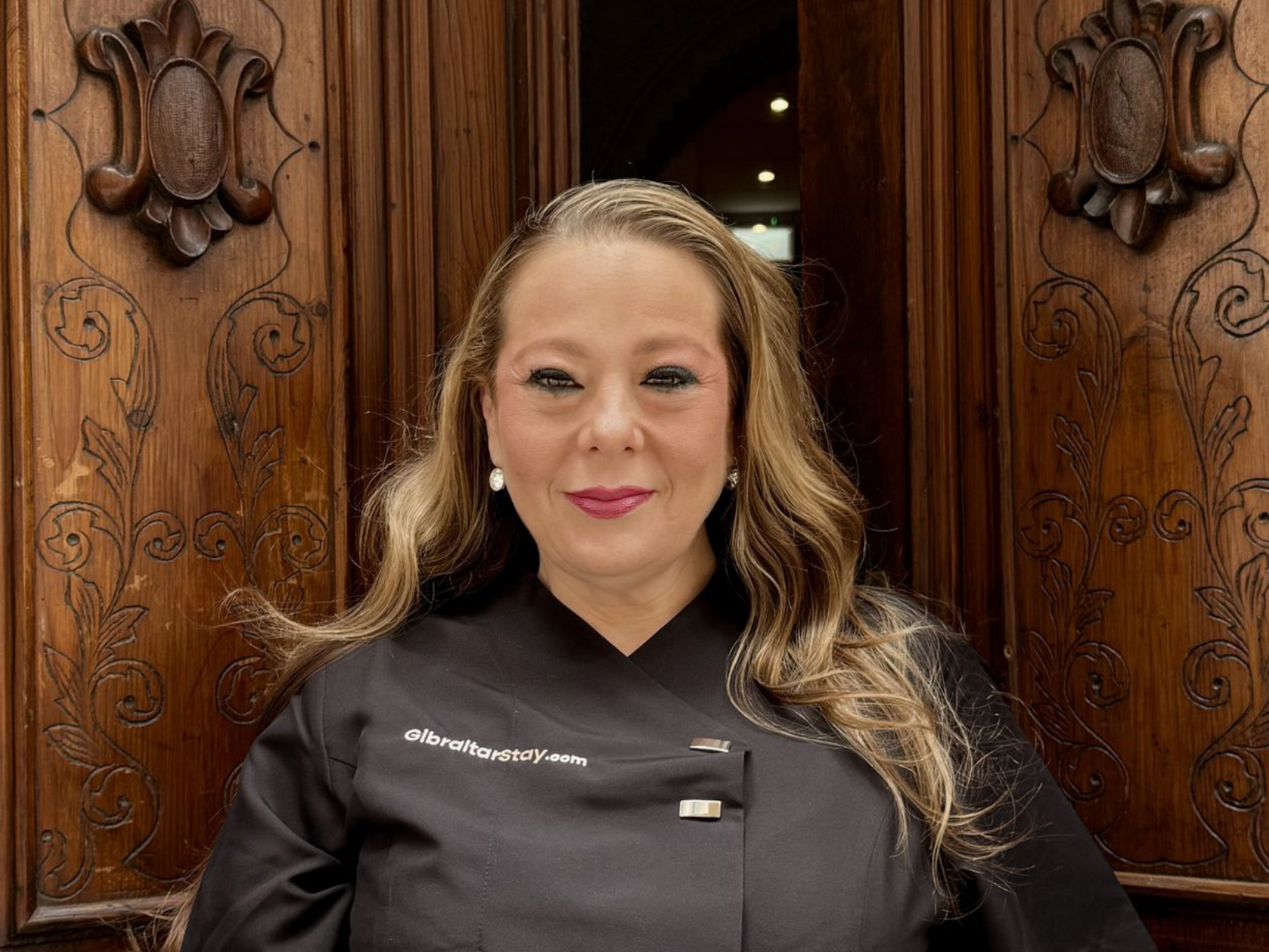 Woman in a black chef coat standing before ornate wooden doors, smiling at the camera.