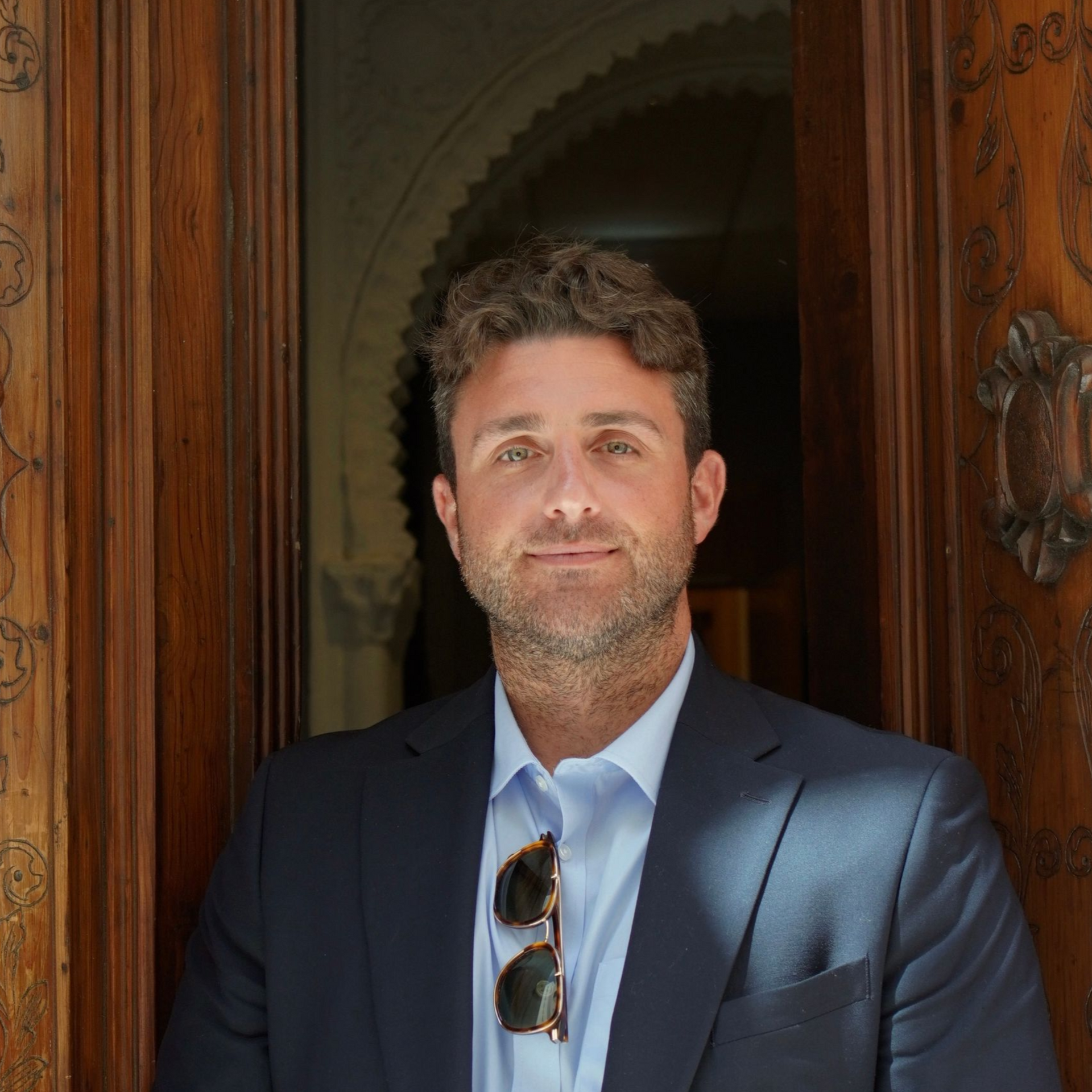 Man in a dark suit stands in a doorway with hands clasped, wearing sunglasses, against a dim interior backdrop.