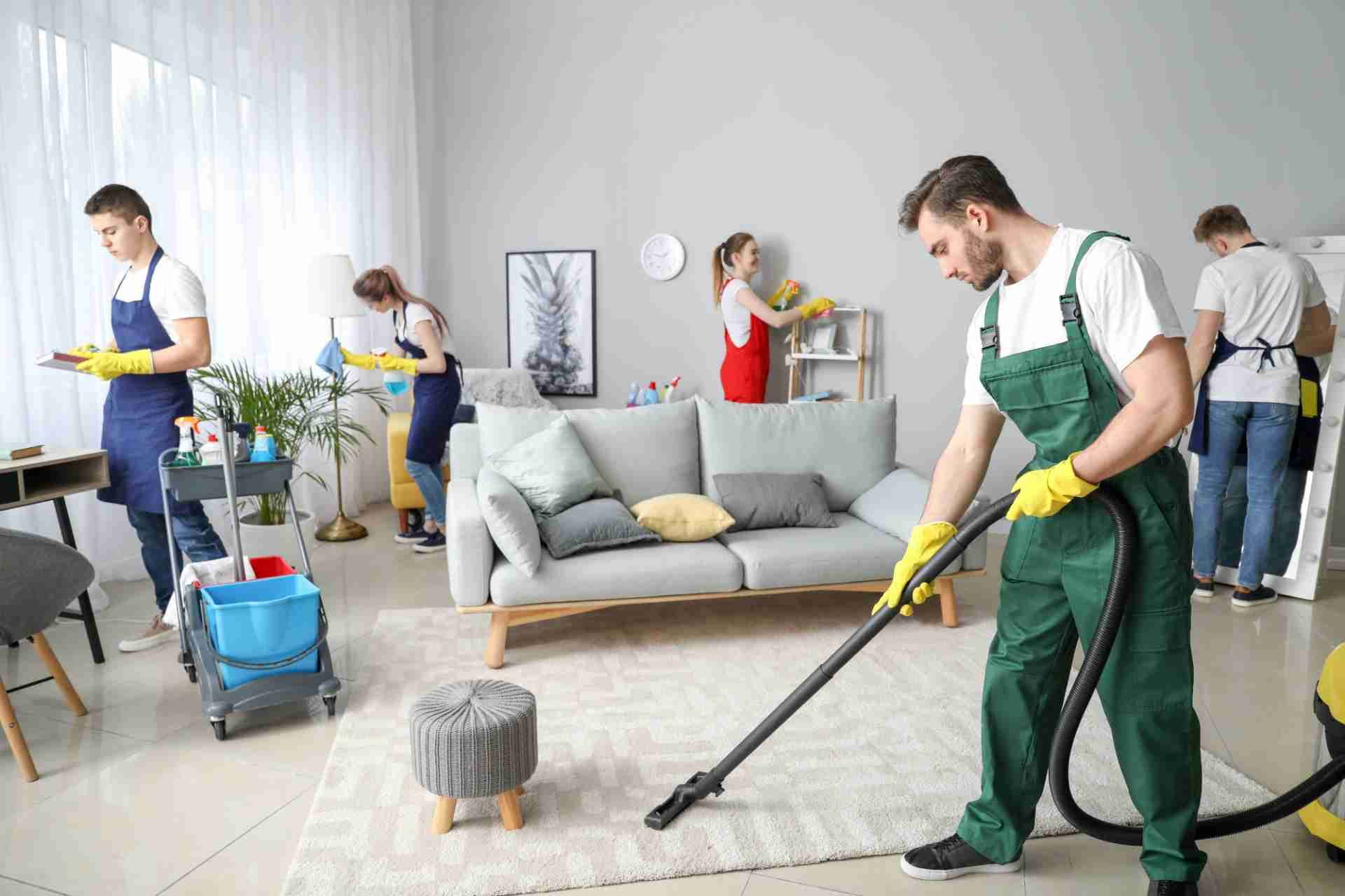 A team of professional cleaners wearing aprons and jumpsuits works in a bright, modern living room.