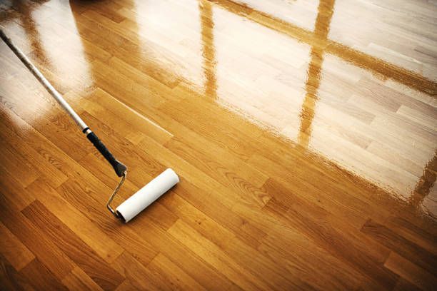 A paint roller applies a glossy finish to a portion of a wooden floor, showing a clear contrast between treated and dry wood.