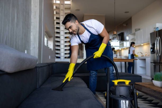 A person wearing a dark apron and yellow gloves vacuums a dark-colored sofa in a bright, open-plan home interior.