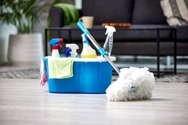 A blue cleaning bucket containing various spray bottles and a cloth, with a white string mop resting against it.
