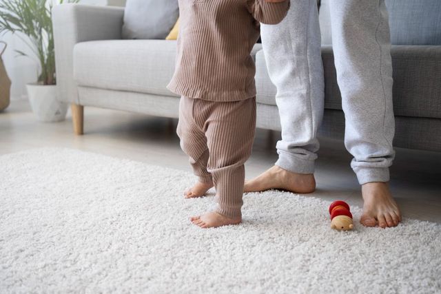 Toddler walking beside an adult in a bright living room, with a small toy on a white rug.