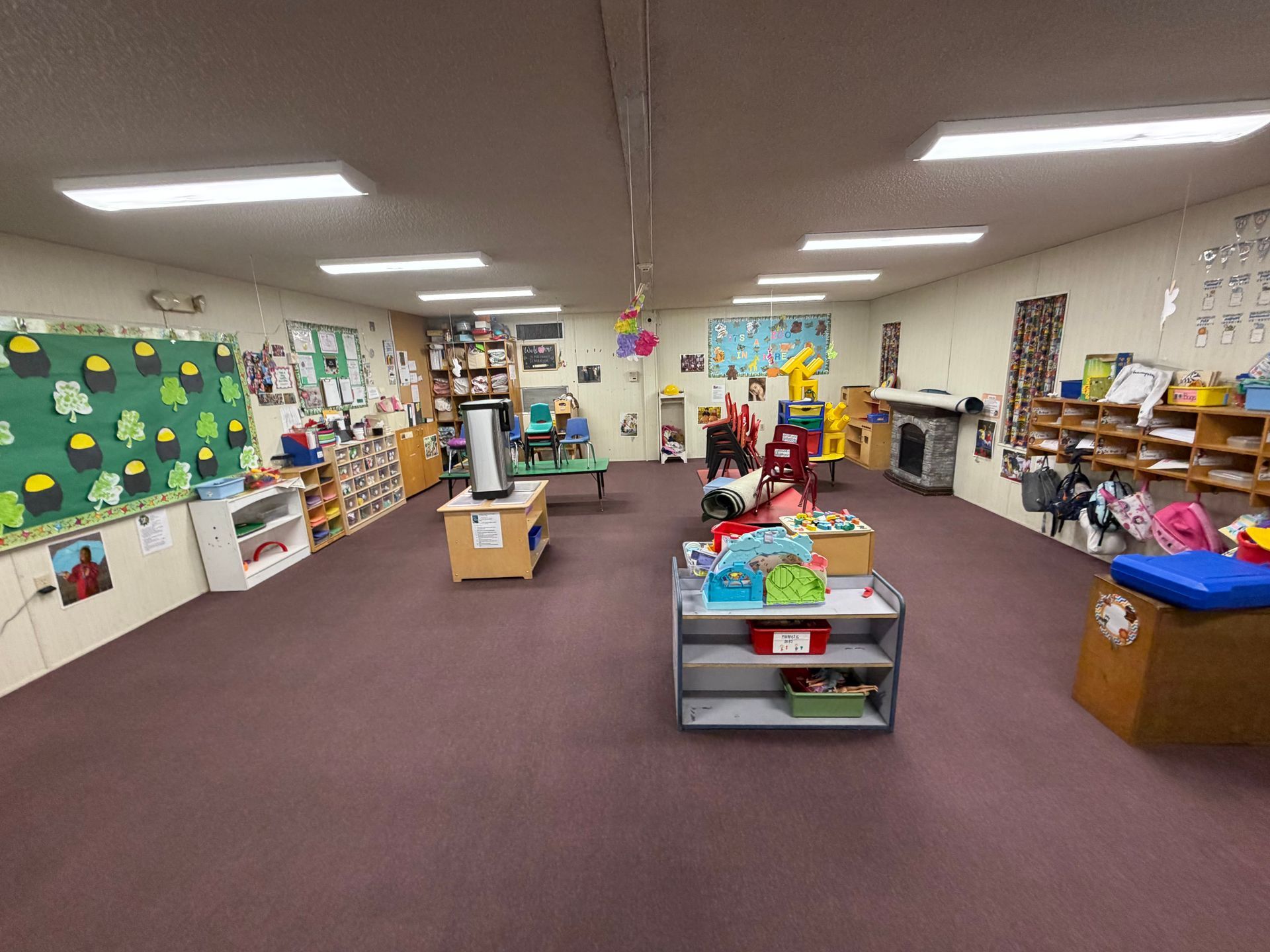 Classroom with carpet, shelves of books and toys, colorful wall displays, and a few children at tables.