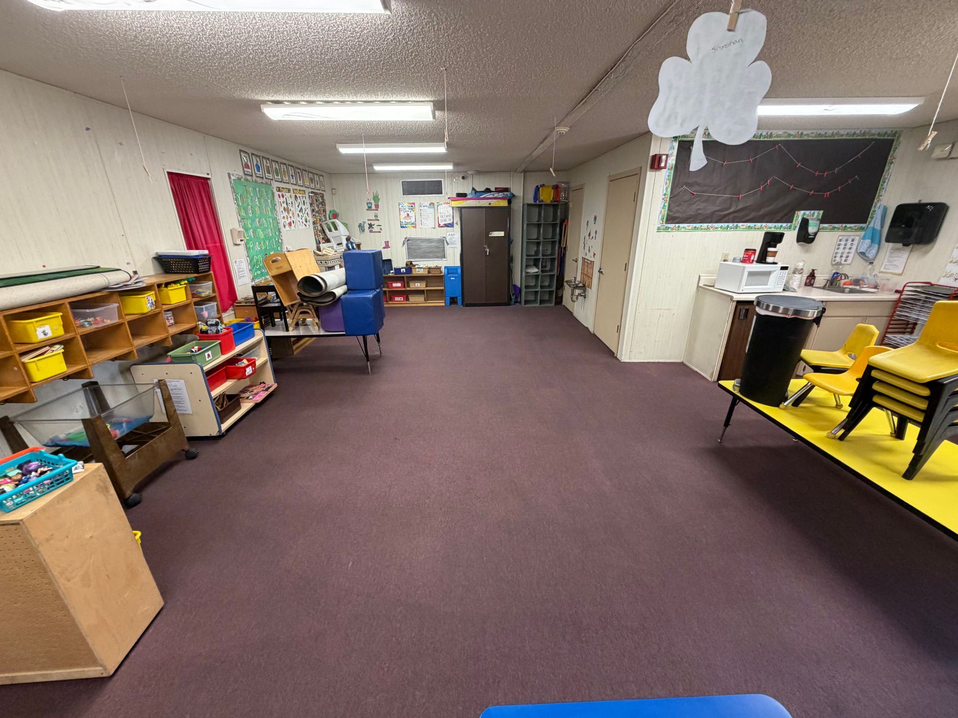 Colorful classroom with shelves, rugs, chairs, and a person arranging items near the back wall.