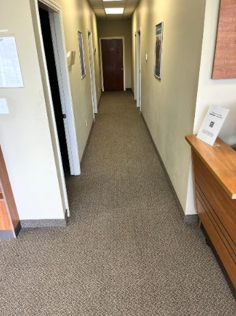 Narrow office hallway with beige walls, carpeted floor, and a wooden cabinet on the right.