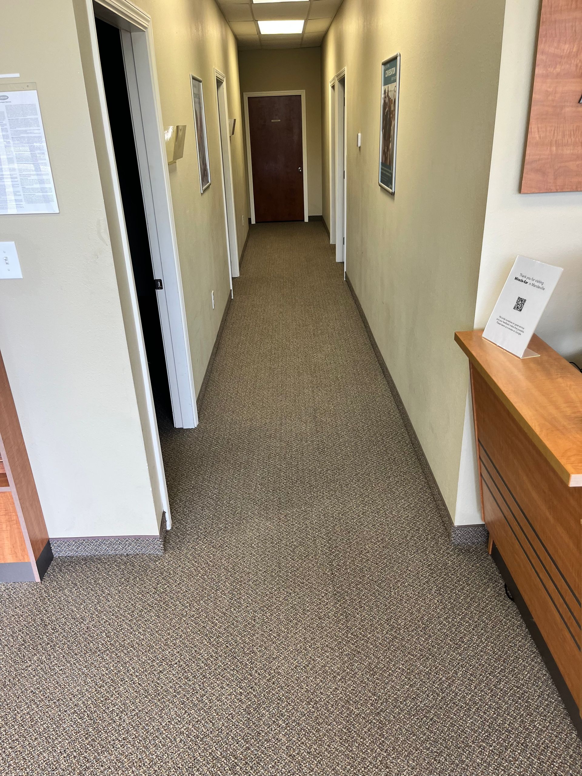 Narrow office hallway with beige walls, patterned carpet, and a wooden cabinet on the right.