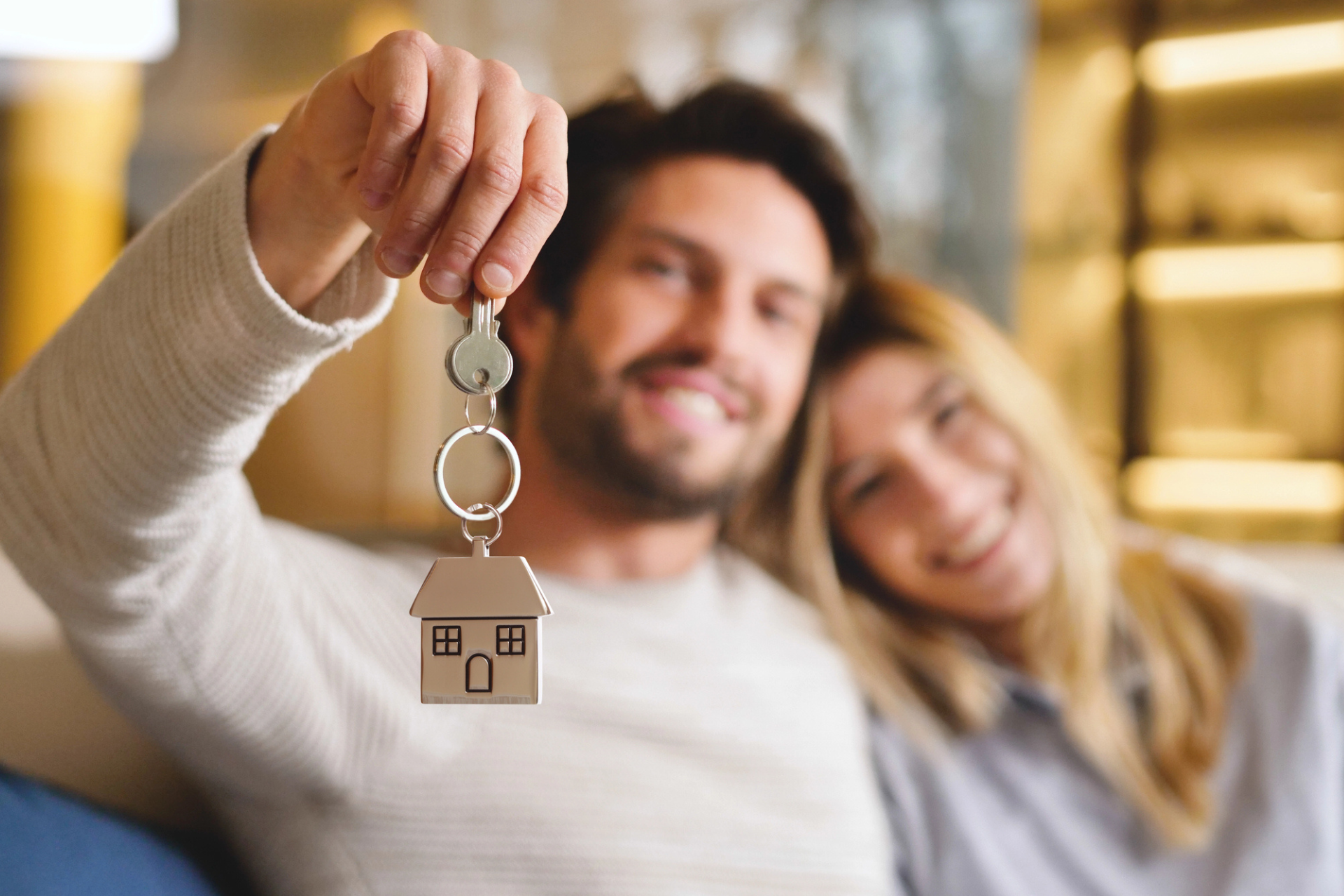 A happy couple sits on a couch, smiling, holding house keys with a small house-shaped keychain.
