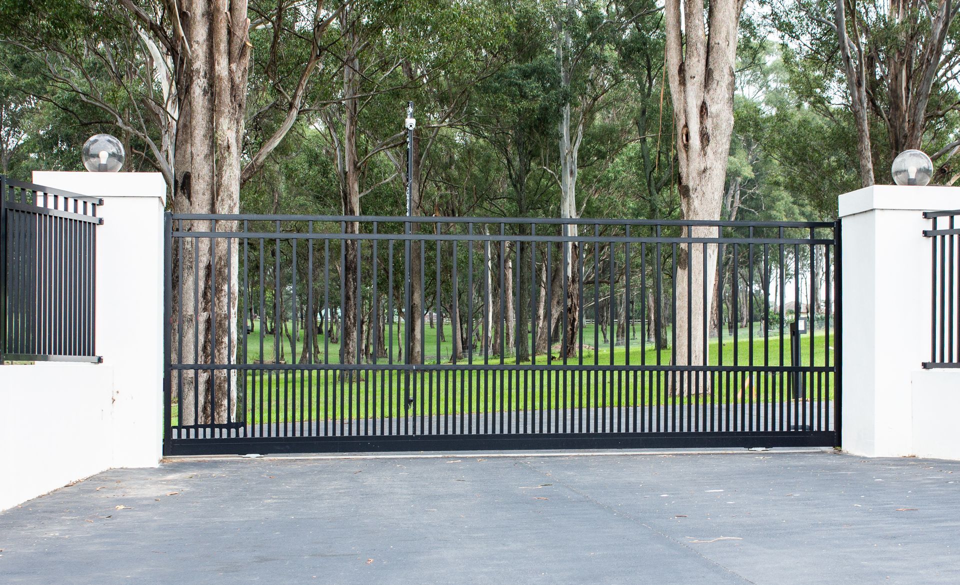 Black metal gate in front of white pillars, leading to trees and a green lawn.