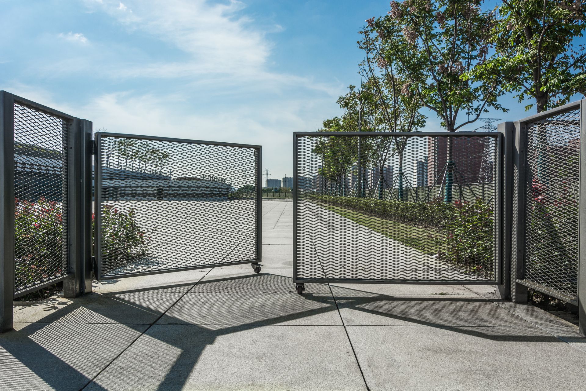 Open, decorative metal gates on a concrete path, leading into a green space under a blue sky.