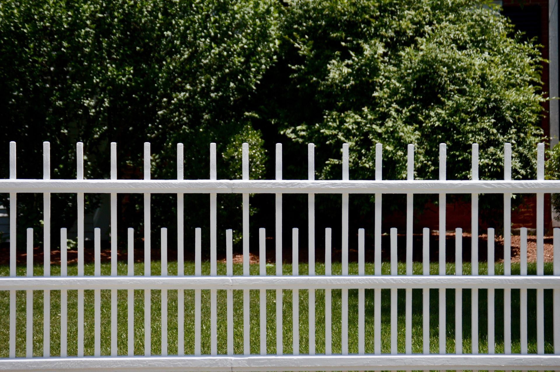White metal fence in front of green lawn and lush green bushes.