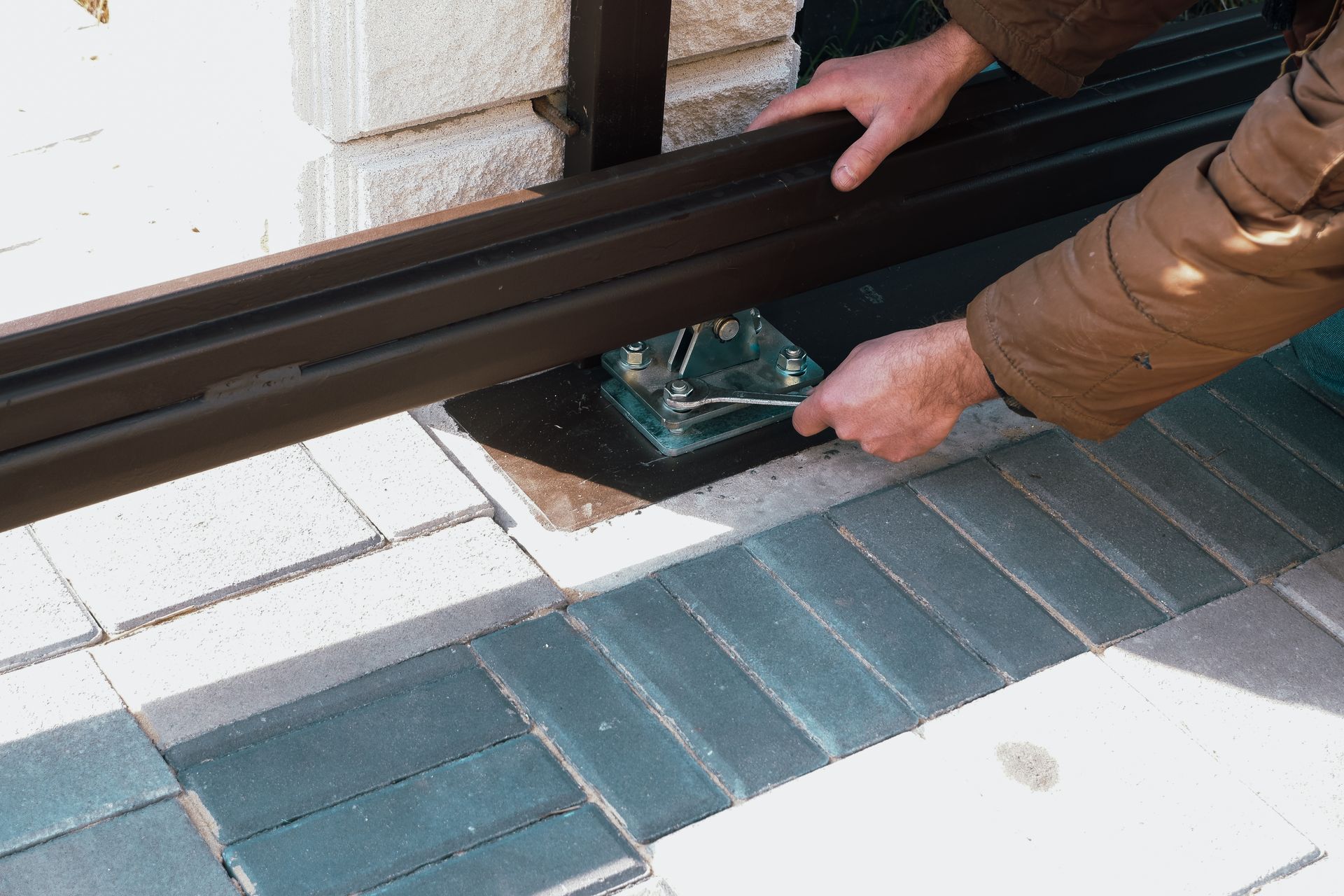 Person tightening bolt on a dark brown metal frame; setting of pavers and brick wall.