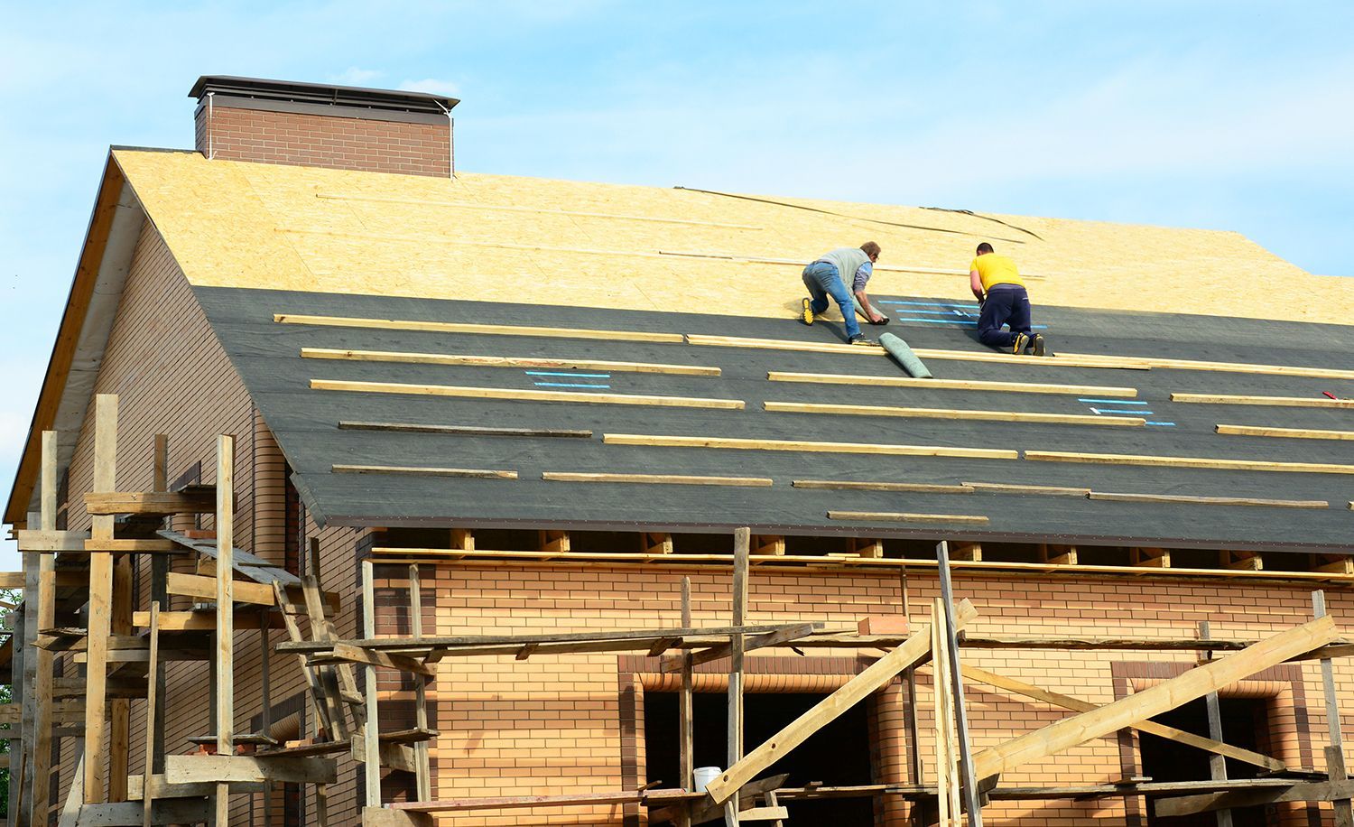 Workers installing shingles on a house roof under construction, with exposed framing below. Workers installing shingles on a house roof under construction, with exposed framing below.
