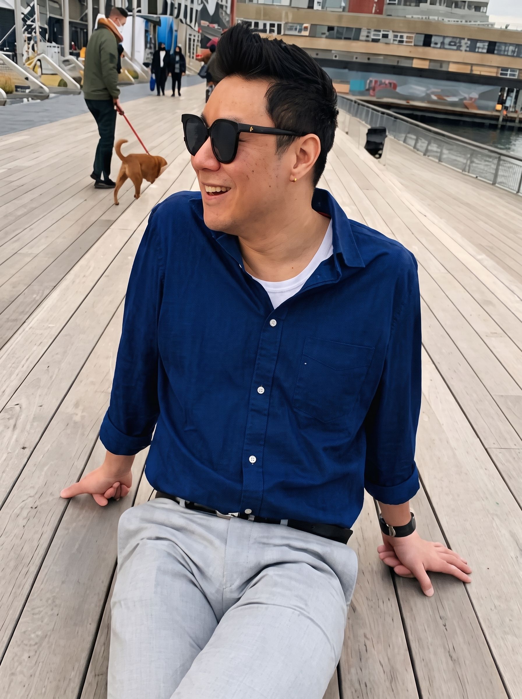Man in blue shirt and sunglasses sitting on a wooden boardwalk, smiling and leaning back outdoors.