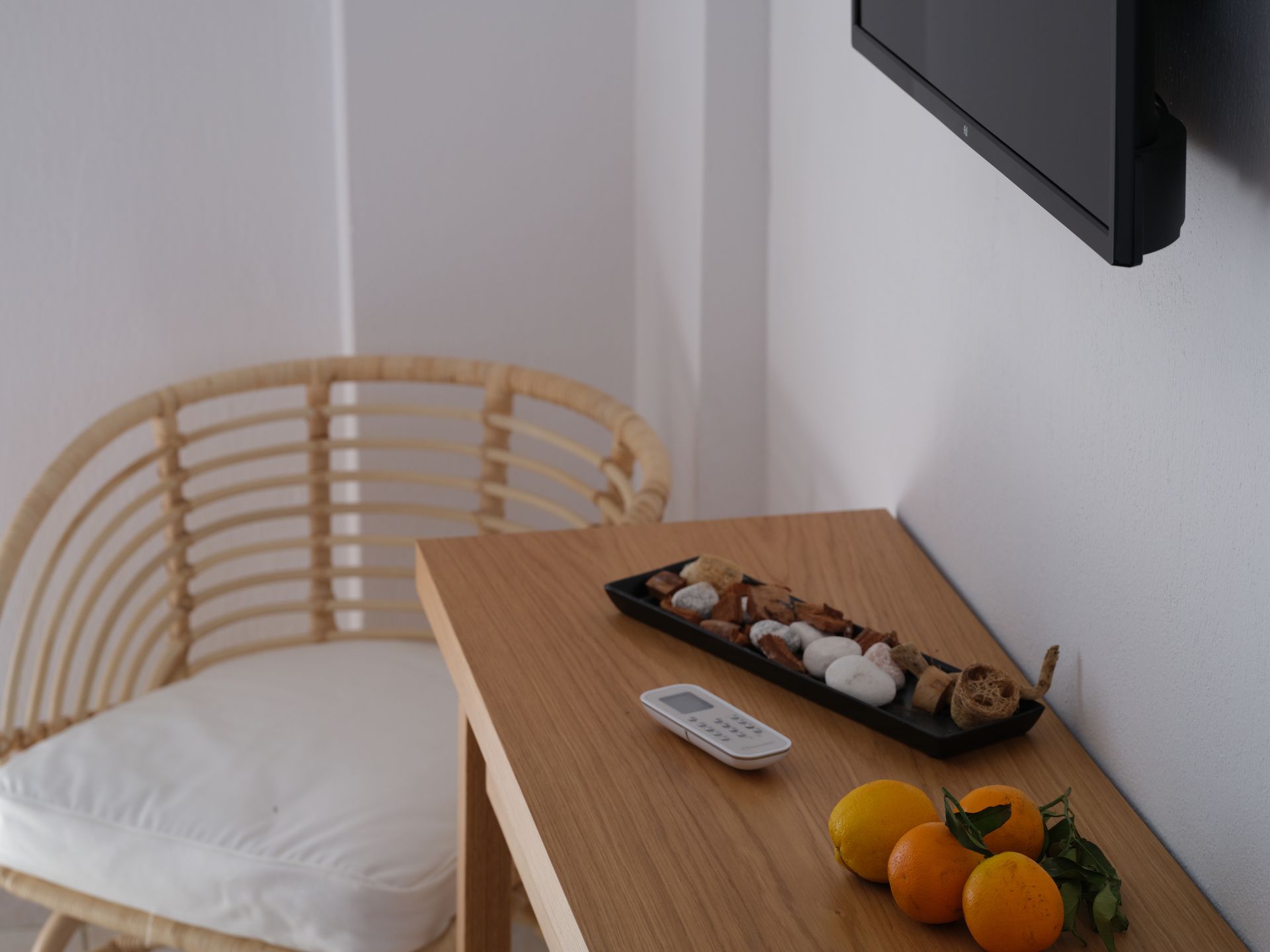 A wicker chair sits next to a wooden table with oranges and a remote control on it