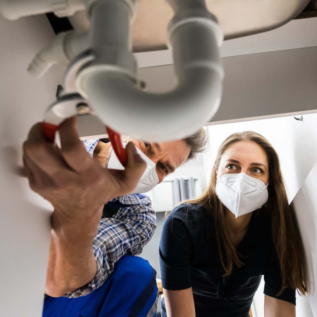 Plumber fixing pipes under a sink as a woman watches, both wearing face masks.