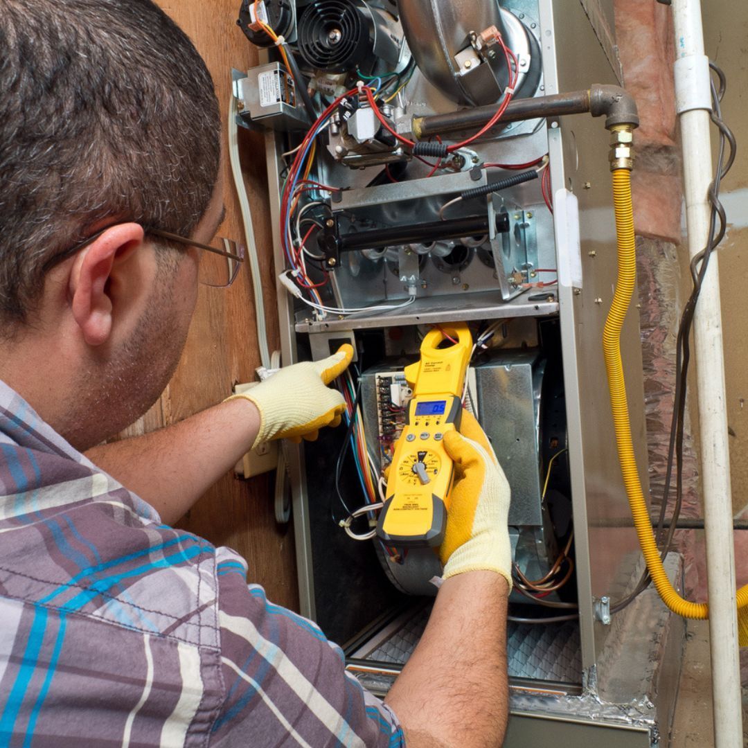 HVAC technician in gloves uses a yellow multimeter to inspect a furnace, indoors.